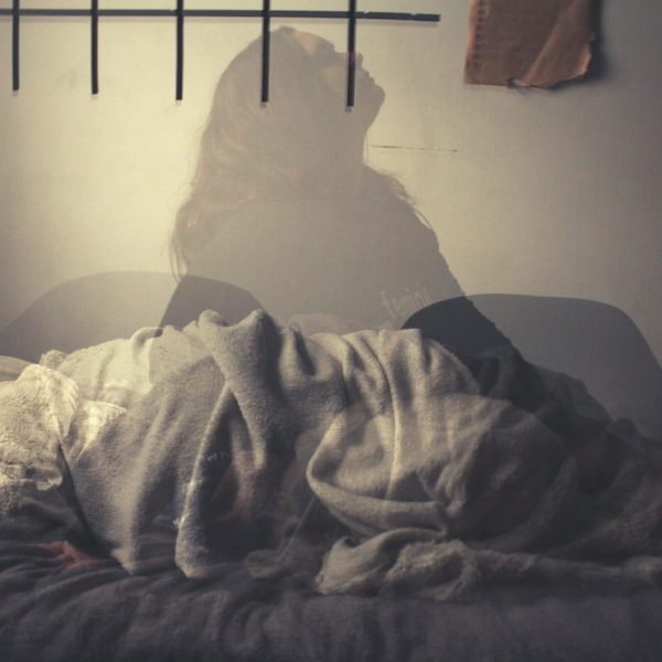 Image of the shape of a person under the covers in bed. Superimposed on this image is a quasi-transparent image of a woman with her head back to depict layers of grief.