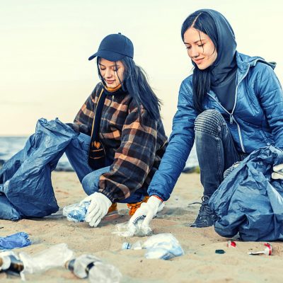 Image of two individuals with trash bags picking up litter off of the beach. Both are dressed in warm clothing and are wearing gloves.